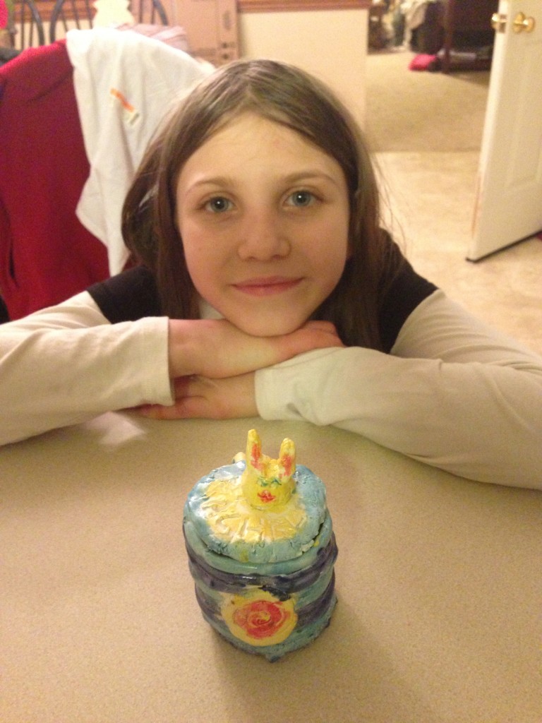 photo of Rachel behind her ceramic art project with a mug that has a bunny sculptured cover. The mug is multi-colored with blues yellow, and rose, and the bunny is mostly yellow.