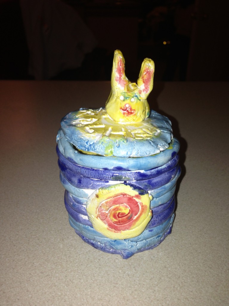 photo of Rachel behind her ceramic art project with a mug that has a bunny sculptured cover.  The mug is multi-colored with blues yellow, and rose, and the bunny is mostly yellow.
